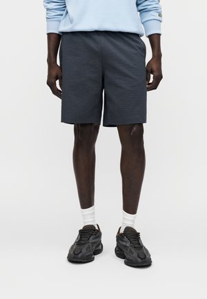 Pantaloncini grigi dalla texture strutturata e vestibilità comoda, con lunghezza a metà coscia. Abbinati a scarpe nere e calzini bianchi alla caviglia.