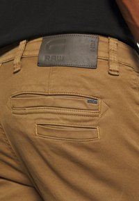 Brown denim pants featuring a textured finish, visible stitching, and a leather label on the waistband with "RAW" branding. Two back pockets are visible.