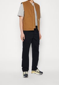 Chaleco acolchado marrón con cremallera dorada, llevado sobre una camiseta gris, combinado con pantalones cargo negros y zapatillas deportivas multicolores.