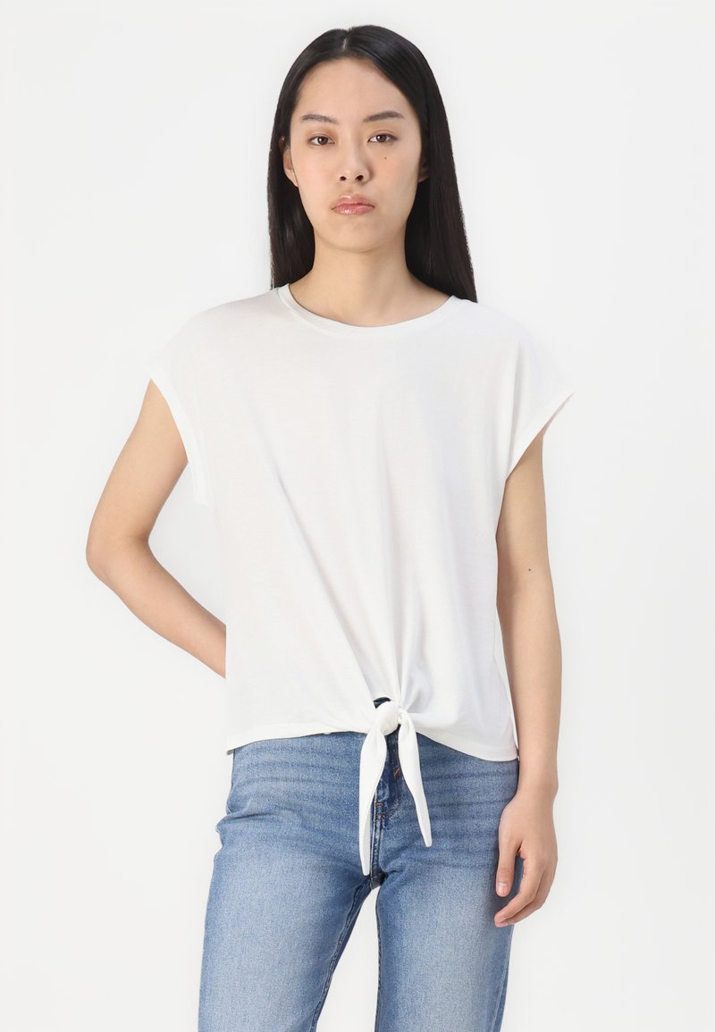 Weißes T-Shirt mit kurzen Ärmeln und Knotendetail an der Taille, aus einem weichen, leichten Stoff gefertigt. Kombiniert mit blauen Jeans.