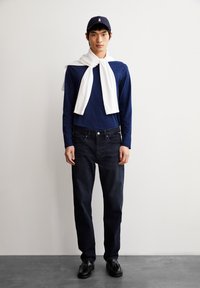 Camisa de manga larga azul marino, suéter blanco colgado sobre los hombros, jeans oscuros y zapatos negros. El modelo está de pie contra un fondo claro.