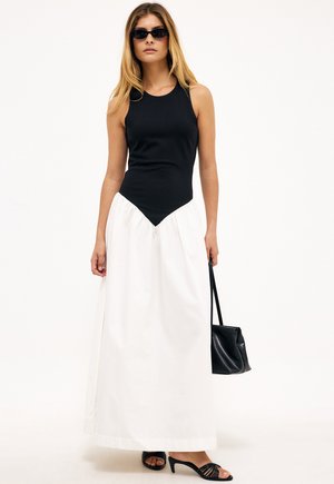 Nainen, jolla on päällään hihaton musta paita ja valkoinen maksihame, mustat korkokengät, aurinkolasit ja musta käsilaukku.