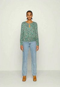 Blusa longa de mangas verdes com estampa floral e decote em forma de chave, combinada com jeans retos azul claro e botins de tornozelo castanho.