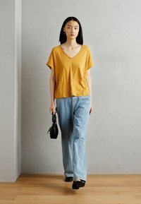 Dzeltena V-veida t-krekla, brīva piegriezuma, saskaņota ar gaiši zilo platajā kājā biksēm. Modelis tur mazu melnu rokassomu. Minimalistisks fons.