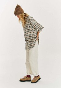 Kobieta w brązowej dzianinowej czapce, szarej koszuli w kratę, kremowych spodniach, czerwonych skarpetkach i beżowych butach do chodzenia po górach, stojąca i patrząca w dół na jednolitym tle.