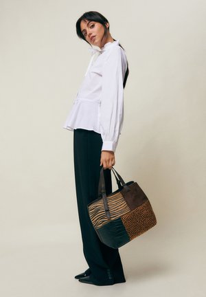 Mujer con blusa blanca y pantalones negros sostiene un bolso grande de parches con estampados de animales sobre un fondo liso.