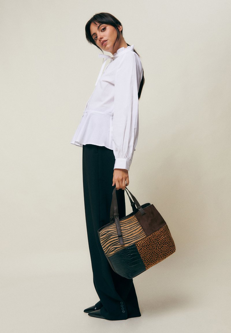 Femme portant un chemisier blanc et un pantalon noir tient un grand sac cabas en patchwork avec des imprimés animaux sur un fond uni.