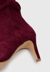 Lähedal pilt burgundia suede stiletto ankle saapast tan värvi kõrge kanna ja valge tausta peal.