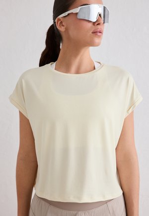 Kobieta nosząca lustrzane sportowe okulary przeciwsłoneczne i luźną, jasnoszarą koszulkę, z włosami związanymi w kucyk.