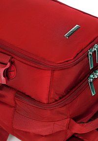Červený látkový batoh s dvěma zipovými kapsami, texturovaným povrchem a kovovými detaily. Obsahuje nastavitelné popruhy a boční poutko.
