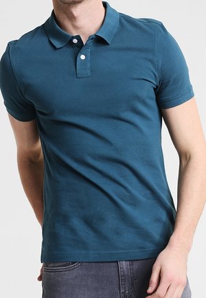 Homme portant un polo uni à manches courtes bleu sarcelle avec deux boutons blancs et un jean gris foncé.