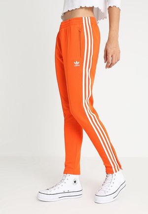 Personne portant un pantalon de survêtement Adidas orange vif avec des bandes blanches et des baskets montantes blanches, debout devant un fond clair uni.