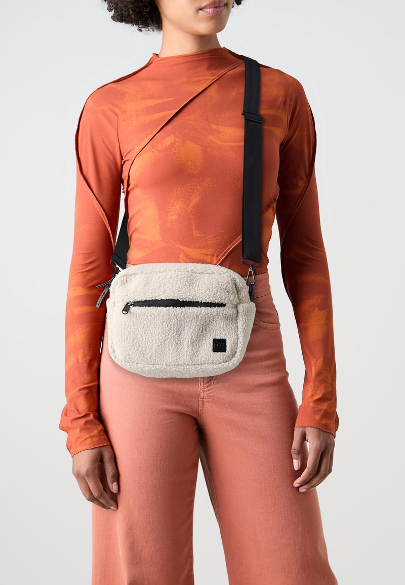 Svetlo béžová crossbody taška z fleecu s čiernym zipsom a nastaviteľným popruhom. Model má na sebe oranžový top s dlhými rukávmi a ružové široké nohavice.