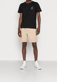 T-shirt en coton noir avec logo blanc sur la poitrine, associé à un short cargo beige. Le modèle porte des baskets montantes blanches.