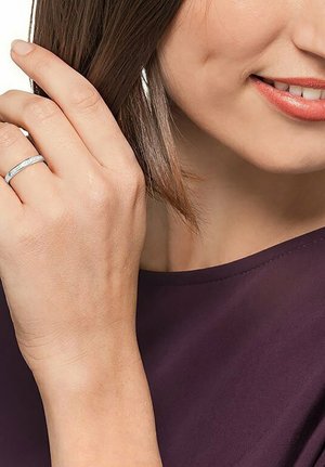 Silberner Ring mit geradem Design, verziert mit kleinen klaren Steinen, getragen an der linken Hand. Hand und Ring vor einem sanften lila Hintergrund.