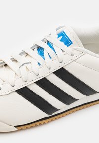 Zapatilla de cuero blanca con tres rayas negras, cordones gris claro y una etiqueta de logo azul. Suela de goma con un patrón en zigzag.