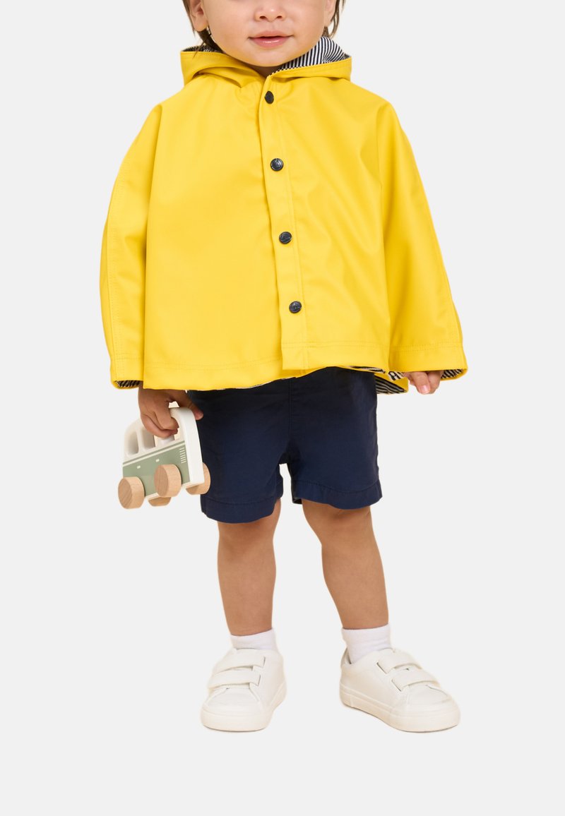 Gelbe wasserdichte Jacke mit Kapuze, schwarzen Knöpfen und gestreiftem Innenfutter; marineblaue Shorts; weiße Turnschuhe; hält ein Holzspielzeugfahrzeug.