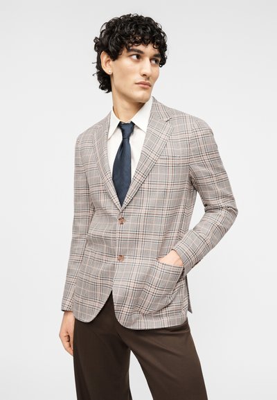 Μπεζ καρό σακάκι με καφέ λεπτομέρειες, μονόπετο σχέδιο, γιακάδες με σχισμές, δύο μπροστινές τσέπες και κλείσιμο με κουμπιά. Συνδυάζεται με λευκό πουκάμισο και μπλε γραβάτα.