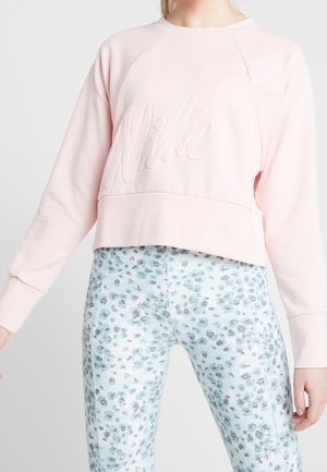 Kobieta ubrana w jasnoróżową bluzę Nike z długim rękawem oraz białe legginsy z wysokim stanem z niebieskimi i szarymi kwiatowymi wzorami.