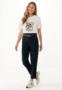 Usměvavá žena s dlouhými vlnitými vlasy, oblečená ve bílém tričku s květinovým potiskem, černých kalhotách s páskem a bílých sportovních botách, stojící na bílém pozadí.