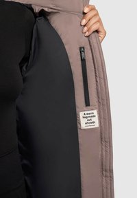 Pufferjack met een taupekleurige buitenkant, zwarte voering, zijrits en een stoffen label met de tekst "Een warme omhelzing gemaakt van stof."