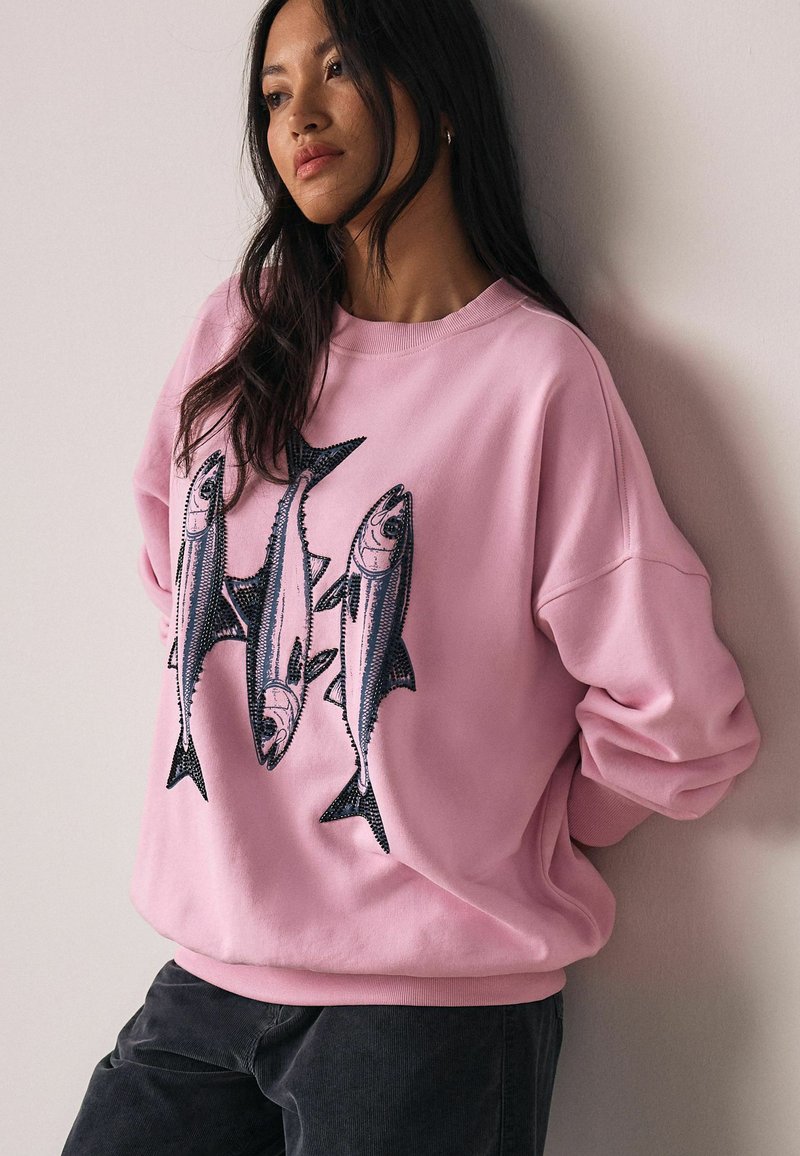 Roze sweatshirt met een relaxte pasvorm, voorzien van zwarte visborduursels aan de voorkant, ronde halslijn en geribbelde manchetten. Zachte textuur.