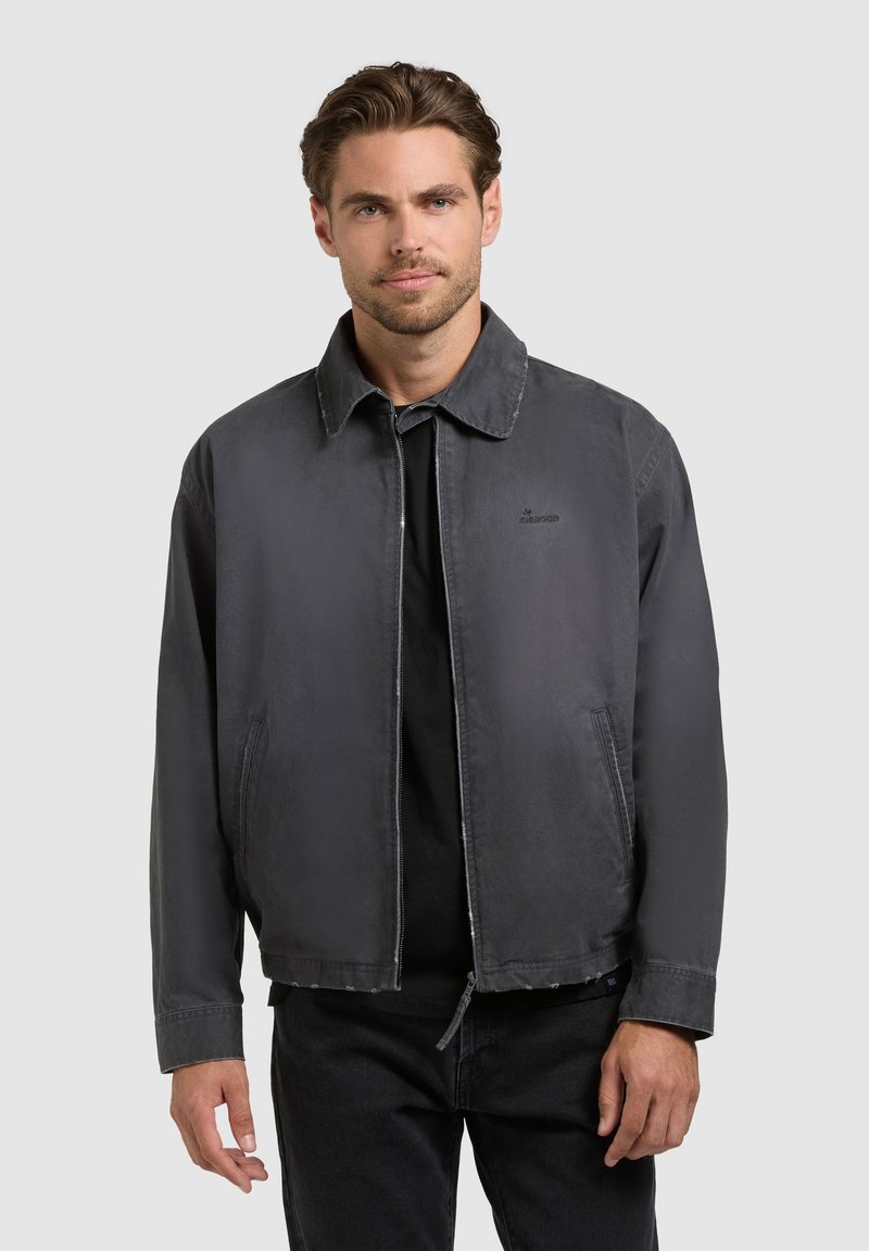Jeune homme aux cheveux bruns courts portant une veste zippée gris foncé sur un t-shirt noir, debout devant un fond clair uni.