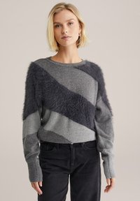 Grauer Pullover mit diagonalen akzentuierten Fleece aus dunkelgrauem Kunstfell, gerippten Bündchen und einem strukturierten Strickdesign. Das Model trägt ihn zu dunklen Hosen.