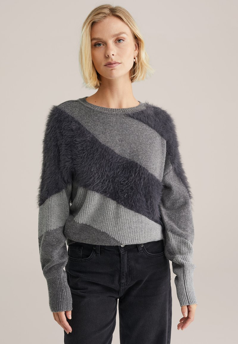 Grauer Pullover mit diagonalen akzentuierten Fleece aus dunkelgrauem Kunstfell, gerippten Bündchen und einem strukturierten Strickdesign. Das Model trägt ihn zu dunklen Hosen.