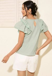 Top in cotone verde menta con maniche corte a volant, bordo scallop, dettaglio con apertura a forma di chiave sul retro, abbinato a pantaloni corti bianchi. Texture e design sottili.