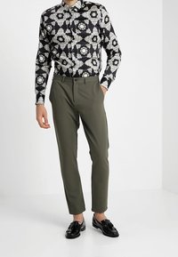 Chemise à motif floral noir et blanc, coupe ajustée, associée à un pantalon tailleur vert olive et des mocassins noirs brillants.