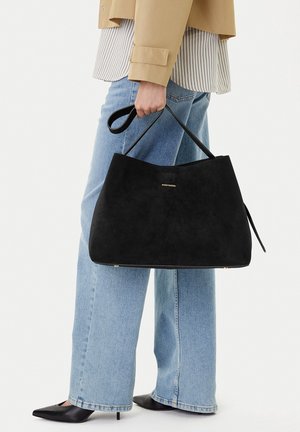 Persona con chaqueta beige, camisa a rayas, vaqueros azul claro, tacones negros puntiagudos, sosteniendo un gran bolso negro con tiras finas.