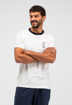 Mann mit dunklem Haar und Bart, trägt weißes T-Shirt mit schwarzem Kragen und Grafik, Arme verschränkt, lächelnd, vor hellem grauem Hintergrund.