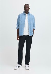 Homme debout avec les mains dans les poches, portant une chemise bleu clair boutonnée par-dessus un t-shirt blanc, un pantalon noir et des baskets blanches avec des rayures noires.