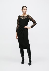 Robe noire avec manches longues en dentelle, corsage ajusté et jupe drapée. Portée avec des bottes noires slim montant jusqu'au genou. Texture lisse avec des accents en dentelle.