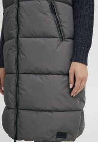 Sivi puffer prsluk s horizontalnim šavovima i dva zip zatvorena džepa. Ima logotip i glatku, sjajnu teksturu tkanine.
