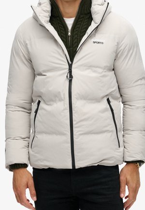 Homme portant une veste matelassée gris clair avec une fermeture éclair à l'avant, par-dessus un pull en maille vert avec un col zippé.