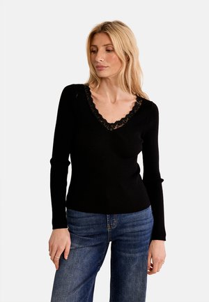 Donna bionda che indossa una maglia nera a maniche lunghe con rifiniture in pizzo sulla scollatura a V e jeans blu, guarda leggermente in basso e di lato.