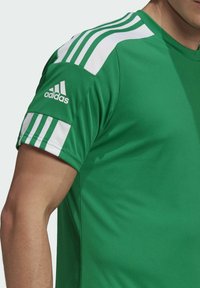 Man i grön Adidas sporttröja med vita ränder på axlar och ärmar, som visar överkropp och arm.