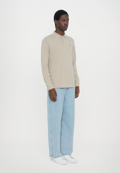 Camisa polo bege de manga longa com colarinho contrastante, combinada com jeans de perna larga azul claro e sapatilhas brancas, de pé contra um fundo simples.