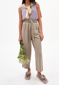 Béžové kalhoty s vysokým pasem a zavazovací šňůrou, doplněné světle fialovým topem bez rukávů a šedými skládacími sandály. Drží zelené květiny.