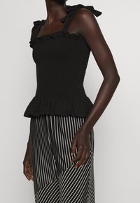 Nainen, joka on pukeutunut mustaan ilmanhihaiseen röyhelötoppia, jossa on nauhakahvat, sekä musta-valkoisiin pystysuoriin raidallisiin housuihin, yksinkertaista taustaa vasten.