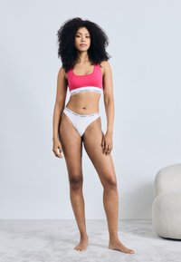 Αθλητικό σουτιέν Calvin Klein σε έντονο ροζ, με λευκή λωρίδα με το λογότυπο, φορεμένο με λευκά ψηλόμεσα σλιπ. Λείο ύφασμα, μινιμαλιστικός σχεδιασμός.