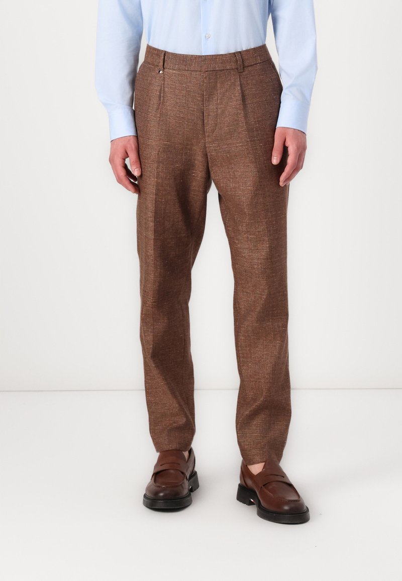 Brown tailored trousers in textured fabric, featuring a pleated front, side pockets, and a relaxed fit. Paired with dark brown shoes.