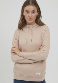 Beige hoodie med dragsko i huvan, känguruficka och ribbade manschetter. Har en liten logotagg vid nederkanten. Mjuk material och avslappnad passform.