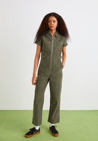 Tutina verde oliva con maniche corte, zip frontale, due tasche sul petto e dettagli con cintura regolabile. Scarpe nere con calzini bianchi.