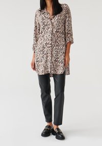 Camicia button-up beige con motivo a foglie nere, maniche lunghe arrotolate. Abbinata a pantaloni di pelle nera e mocassini neri.