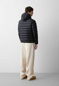 Giacca a vento nera con cappuccio, design trapuntato e dettaglio con logo. Indossata con pantaloni larghi di colore chiaro e calzature beige.