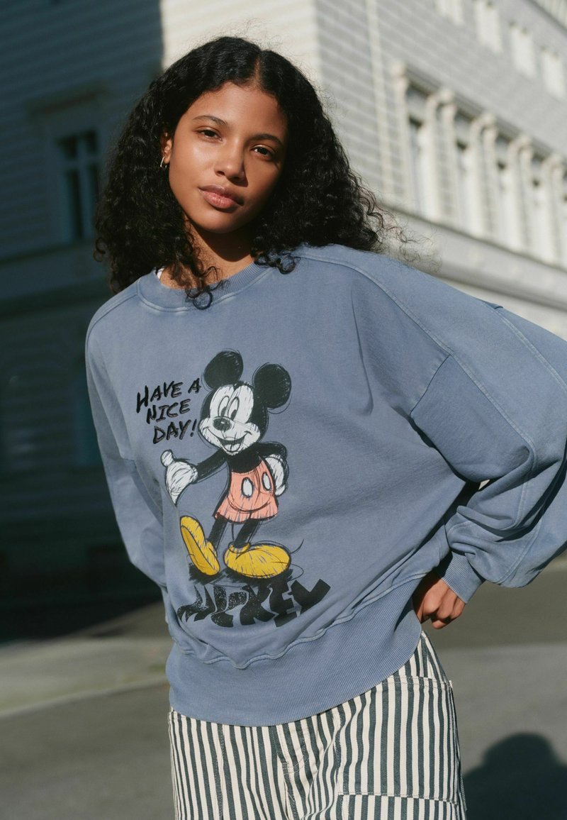 Jeune femme aux cheveux bouclés portant un sweat-shirt bleu Mickey Mouse avec l'inscription « Passe une bonne journée ! » et un pantalon rayé, debout en plein air.