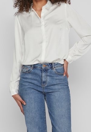 Frau trägt ein weißes langärmeliges Hemd mit Knöpfen, das in hoch taillierte blaue Jeans gesteckt ist, mit einer Hand in der Hüfte und einer entspannt.